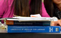 Two textbooks sit flat on a desk, spines out, with a stack of papers on top. The papers mostly cover the spine of the top book. The spine of the bottom book is blue with white text that reads Linguistics of American Sign Language Fifth Edition.